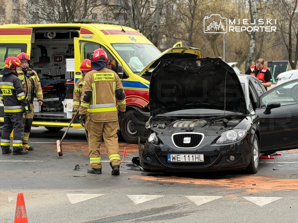 Grupa strażaków i ratowników medycznych pracuje przy wypadku drogowym, obok uszkodzonego czarnego samochodu.