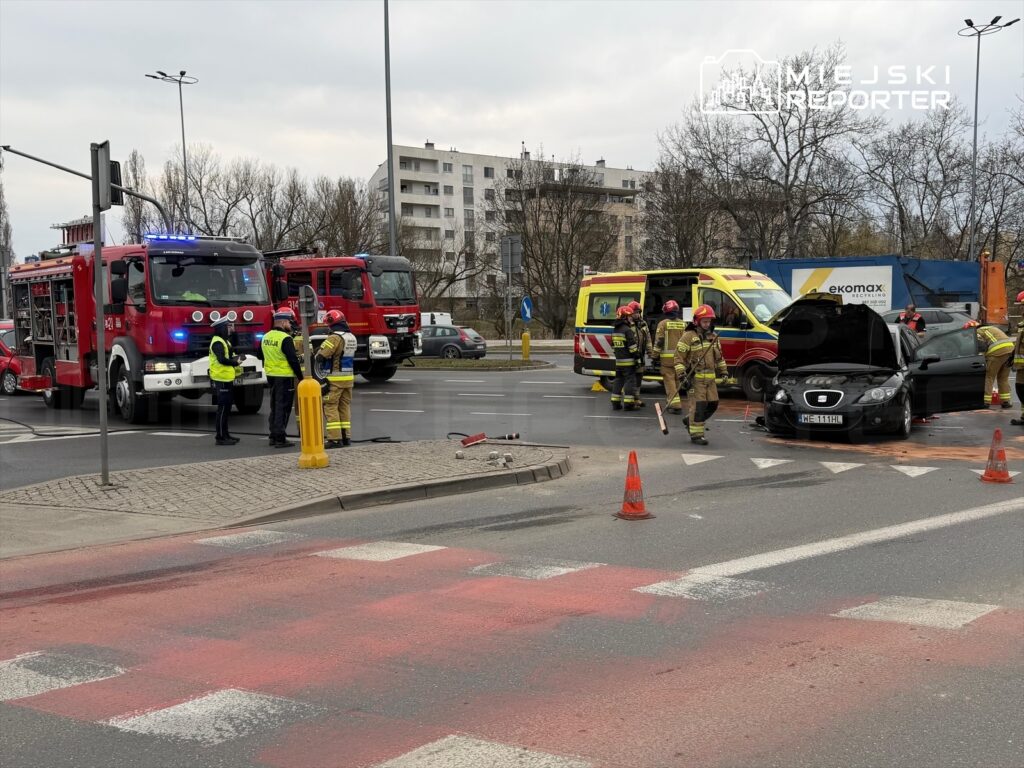 Na skrzyżowaniu, strażacy i policjanci pracują przy rozbitym samochodzie, obok stoją wozu strażackie i karetka.