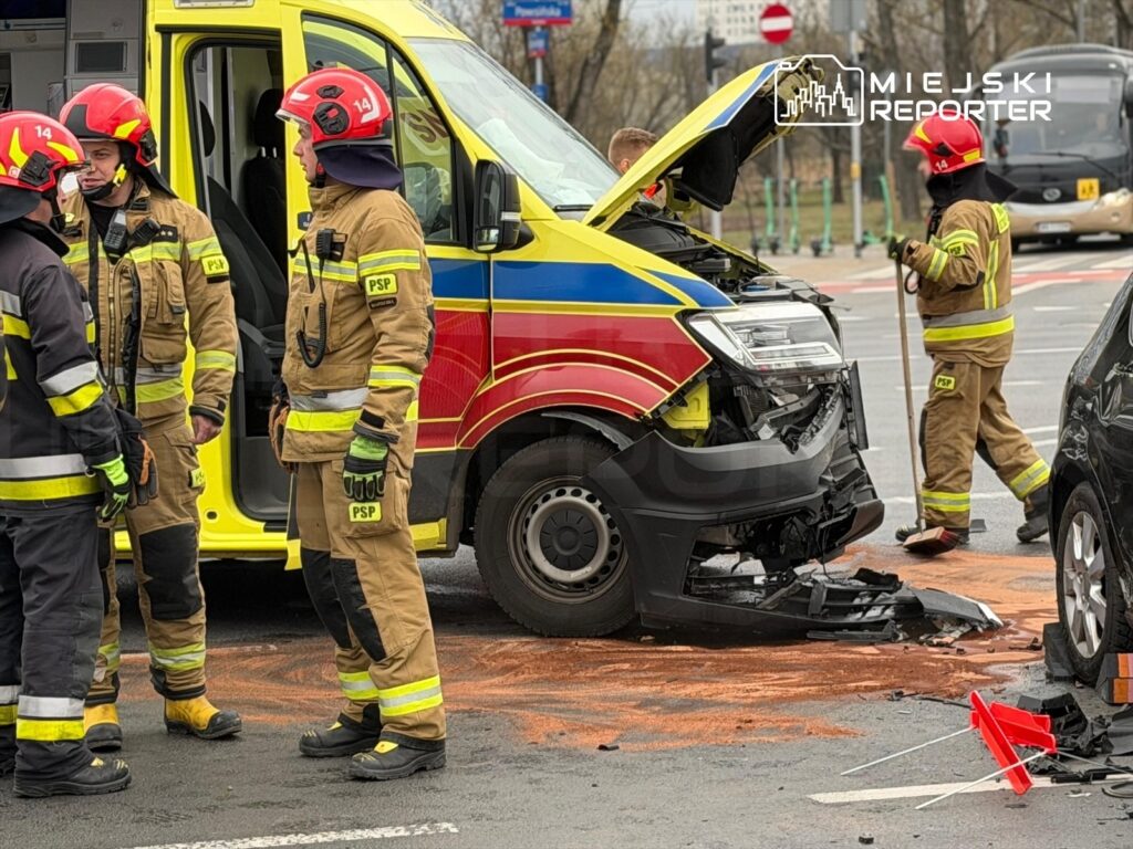 Czterech strażaków w mundurach omawia sytuację przy uszkodzonym pojeździe ratunkowym na skrzyżowaniu.