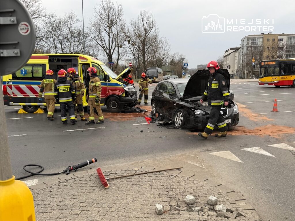 Grupa strażaków pracuje nad usunięciem skutków wypadku drogowego, obok uszkodzonego samochodu osobowego.