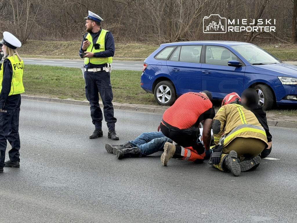 Trzech ratowników udziela pomocy mężczyźnie leżącemu na jezdni, obok nich stoją policjanci. W tle widoczny niebieski samochód.