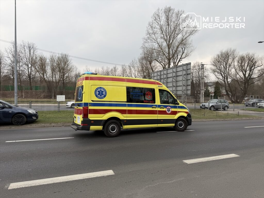 Karetka pogotowia z niebieskimi światłami przejeżdża obok ruchliwej ulicy w miejskim otoczeniu.