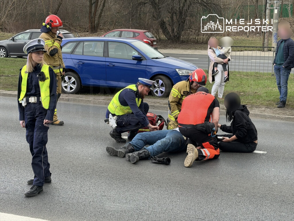 Grupa ratowników oraz policjantka udzielają pomocy poszkodowanemu mężczyźnie na jezdni.