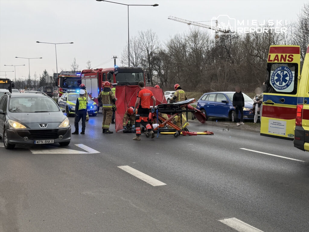 Ratownicy medyczni i strażacy udzielają pomocy poszkodowanemu na drodze, obok przewróconego pojazdu.