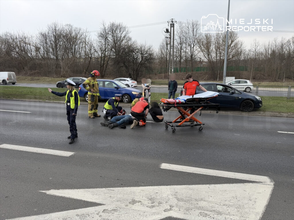 Grupa ratowników medycznych udziela pomocy poszkodowanemu na jezdni, obok stoi policjant kierujący ruchem.