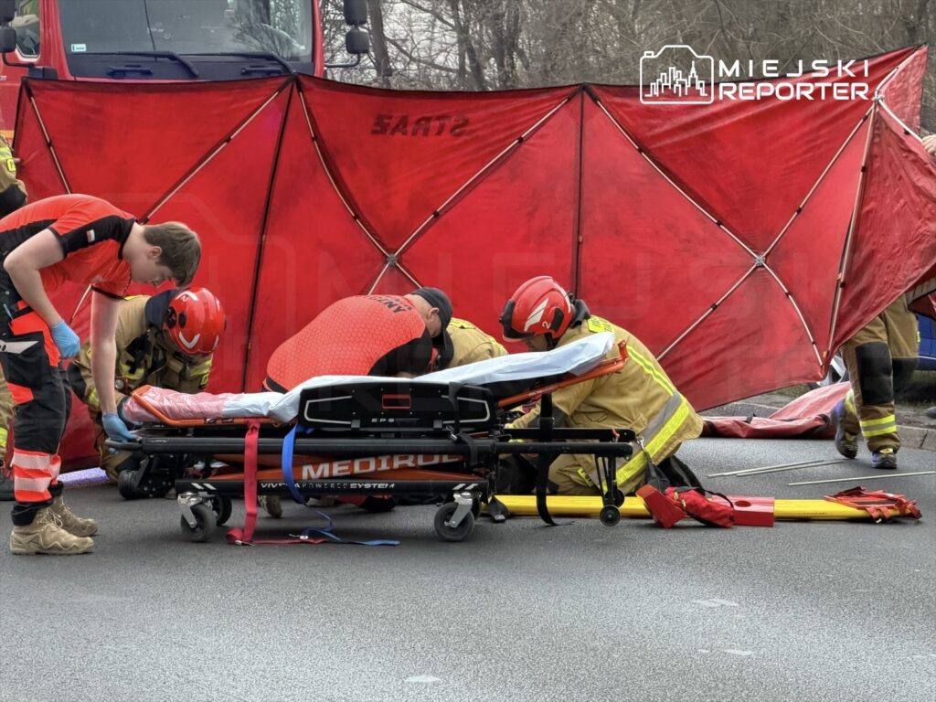 Ratownicy medyczni i strażacy pracują nad poszkodowanym na noszach, otoczeni czerwoną osłoną na drodze.