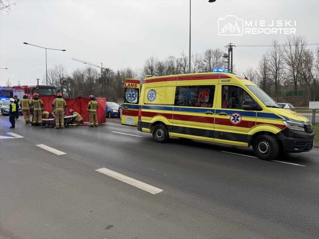 Ratownicy medyczni udzielają pomocy poszkodowanemu na jezdni, obok ambulansu i wozu strażackiego.