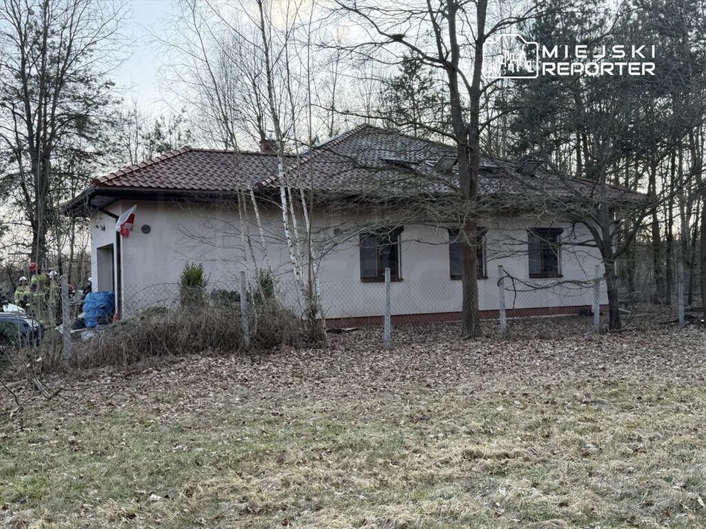 Grupa strażaków w mundurach przy budynku mieszkalnym z flagą Polski na elewacji, otoczonym drzewami.