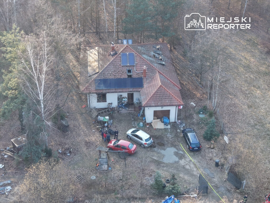 Grupa osób rozmawia obok domu z czerwoną i białą osobówką, w otoczeniu leśnym. W tle widoczne są panele słoneczne na dachu.