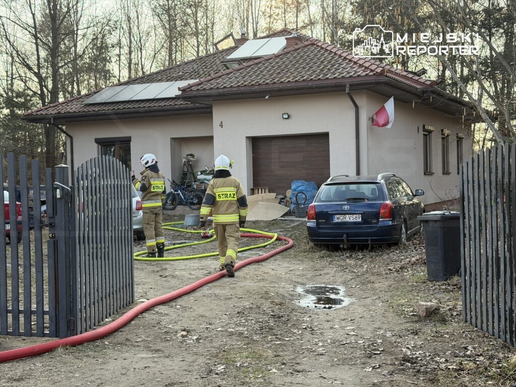Dwaj strażacy w mundurach, z wężami gaśniczymi, pracują przed domem z czerwoną flagą na elewacji.