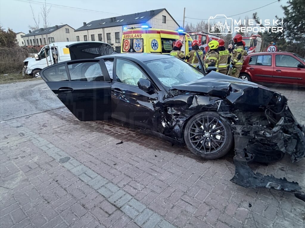 Czarny samochód osobowy z uszkodzoną karoserią stoi na drodze, obok grupy strażaków i karetki.
