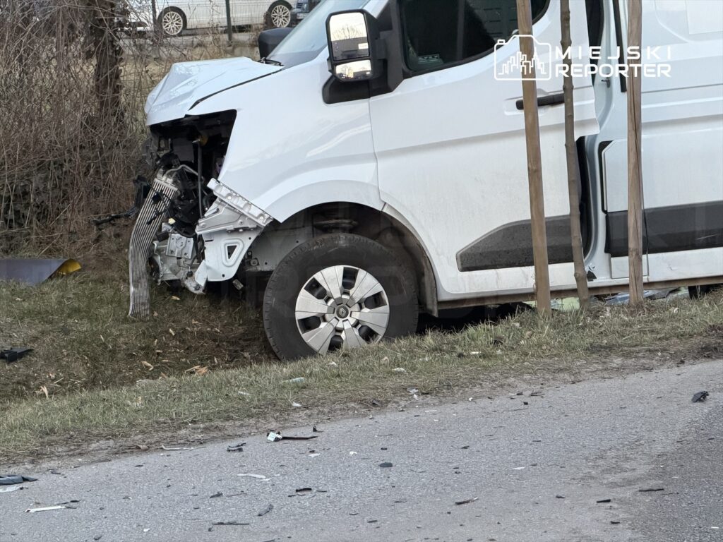 Uszkodzony biały furgon z uszkodzoną przednią częścią stoi na poboczu drogi, otoczony fragmentami wraku.
