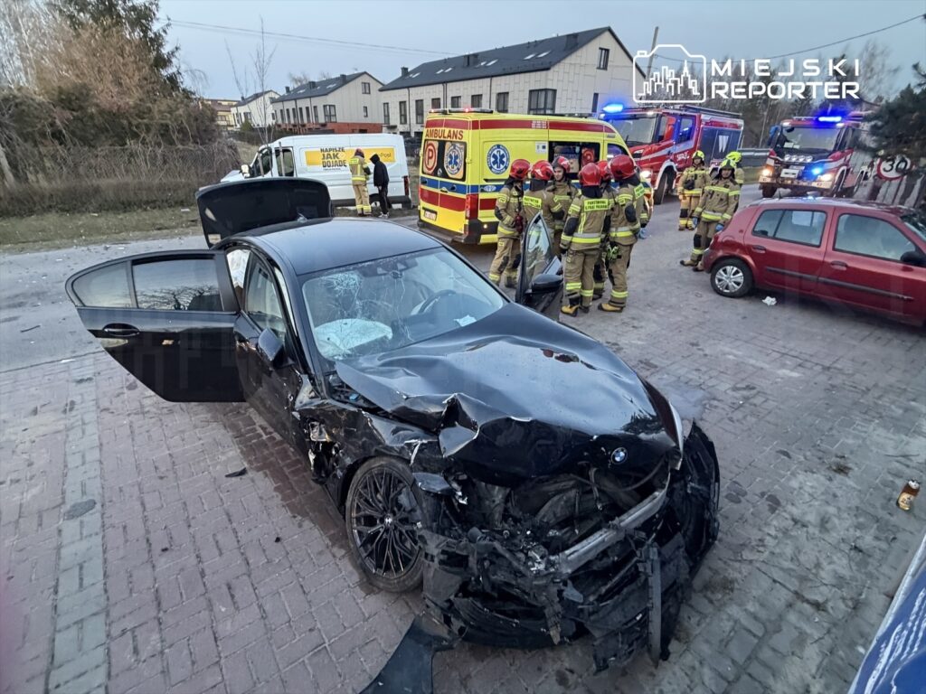 Uszkodzony czarny samochód osobowy stoi na chodniku, obok grupy strażaków i pojazdów ratunkowych.