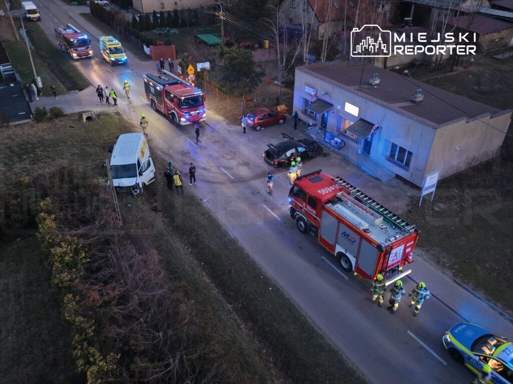 Na drodze stoją strażacy i policjanci, obok uszkodzony samochód dostawczy oraz dwa pojazdy ratunkowe.