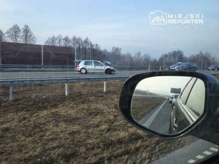 Zniszczony samochód osobowy zatrzymany na poboczu drogi, w tle widoczne inne pojazdy w ruchu.