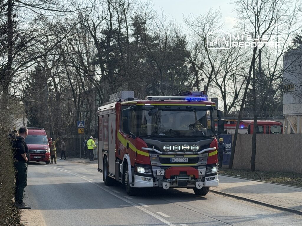 Czerwony wóz strażacki z migającymi światłami przejeżdża obok grupy ludzi na ulicy w otoczeniu drzew.