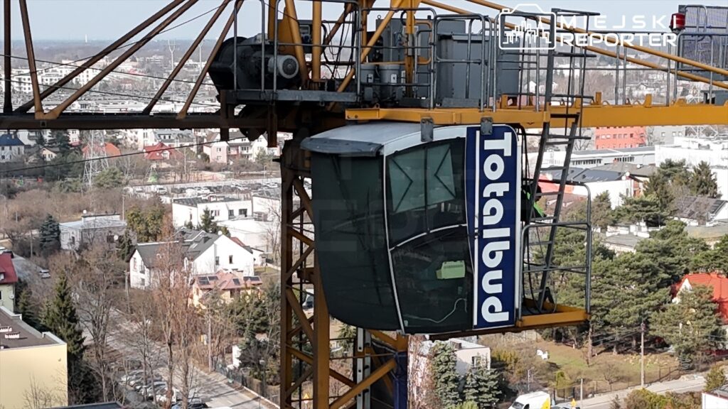 Żuraw budowlany z kabiną operatora, z widokiem na osiedle domów jednorodzinnych w tle.