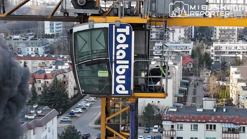 Konstruktor w żółtej kamizelce pracuje w kabinie dźwigu budowlanego, z widokiem na osiedle mieszkaniowe.