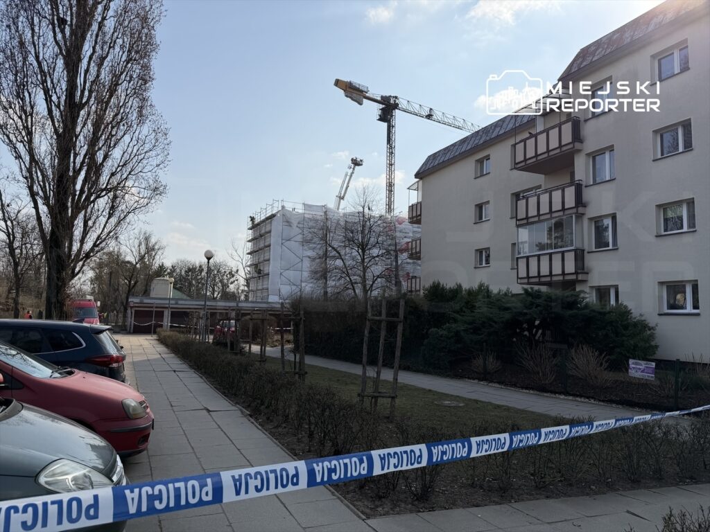 W tle widać budynek w trakcie remontu, z rusztowaniami i osłonięty folią, a przed nim zaparkowane samochody oraz taśma policyjna.