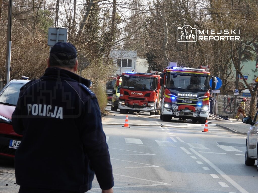 Policjant w mundurze obserwujący akcję ratunkową, w tle dwa wozy strażackie z włączonymi sygnałami świetlnymi.