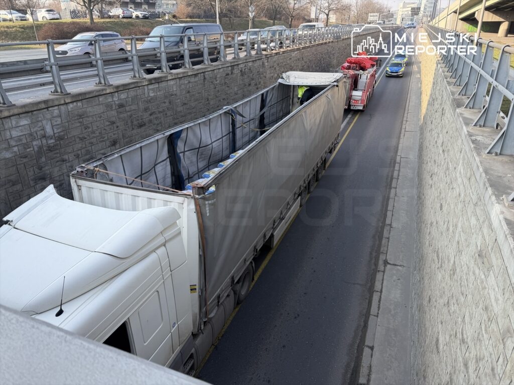Ciężarówka z podniesioną plandeką stoi w wąskim tunelu drogowym, obok przejeżdża pojazd służb ratunkowych.