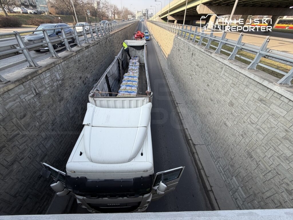 Ciężarówka załadowana workami stoi w wąskim tunelu, obok niej inny pojazd oraz ruch uliczny na górze.
