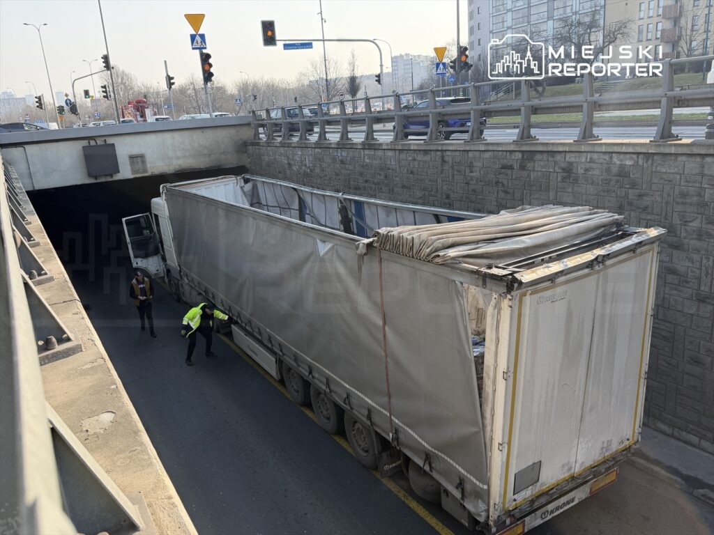 Kierowca ciężarówki z otwartą klapą załadunkową, rozmawia z policjantem przy wjeździe do tunelu.