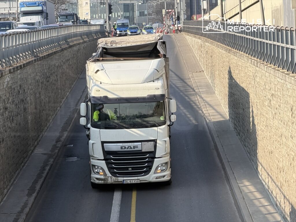 Ciężarówka marki DAF z otwartą naczepą jedzie w wąskim tunelu drogowym, obok dostrzegalne inne pojazdy.
