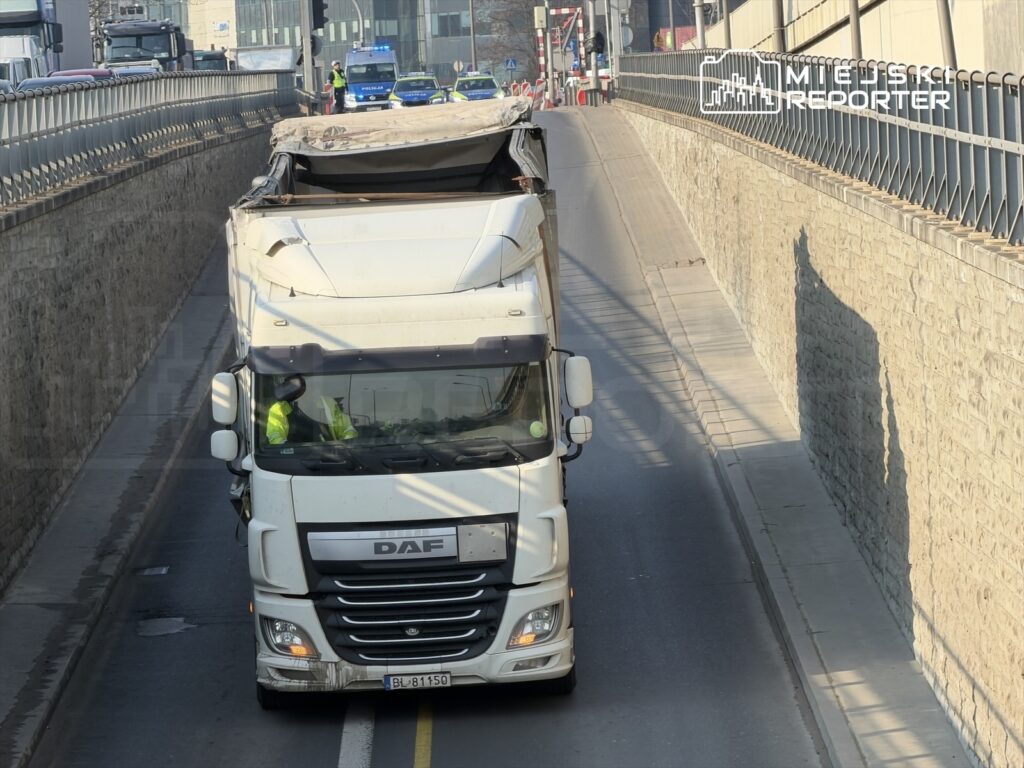Ciężarówka marki DAF z kierowcą w żółtej kamizelce jedzie w dół po wąskiej ulicy, obok kamiennego muru.