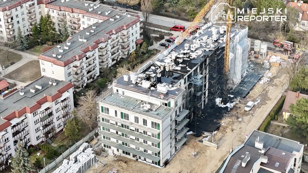 Nowoczesny budynek w budowie z częściowo odsłoniętą elewacją, otoczony przez osiedle mieszkalne i plac budowy.