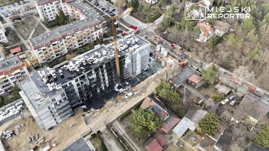 Nowoczesny budynek w trakcie budowy z widoczną dźwigiem, otoczony przez inne budynki mieszkalne oraz pojazdy straży pożarnej.