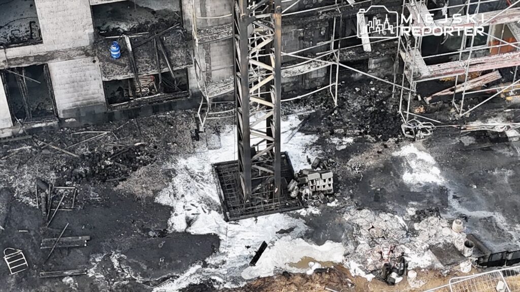 Zniszczony teren budowy z płonącymi resztkami materiałów budowlanych i dużą dźwigiem w centrum.