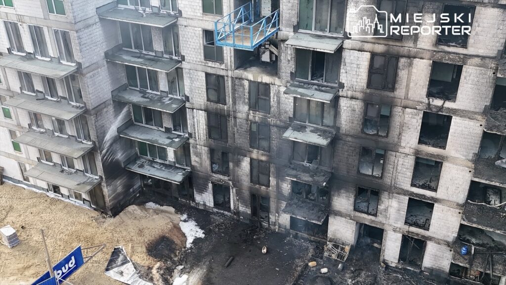 Spalony budynek mieszkalny z uszkodzonymi oknami i czarnymi śladami po ogniu w otoczeniu placu budowy.