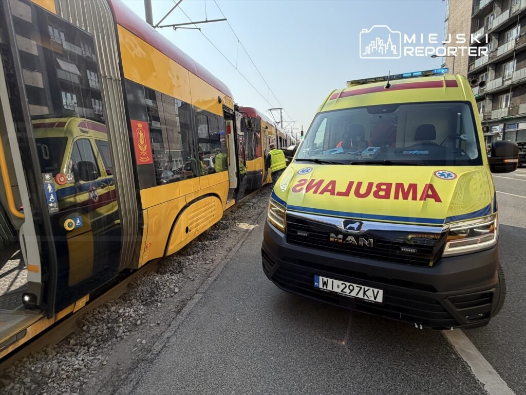 Ambulans z widocznym napisem "AMBULANS" stoi obok żółtej tramwajowej linii na miejskiej ulicy.