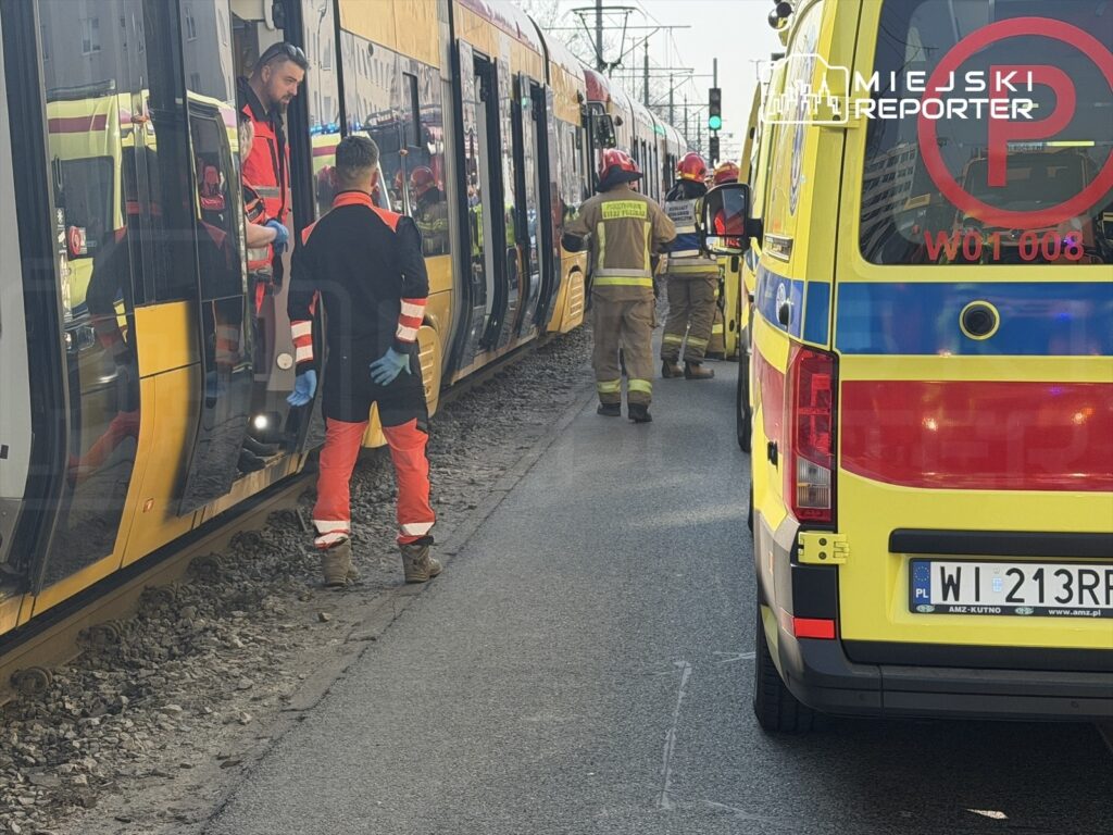Ratownicy w mundurach rozmawiają z pasażerami w tramwaju, obok stoi karetka pogotowia.