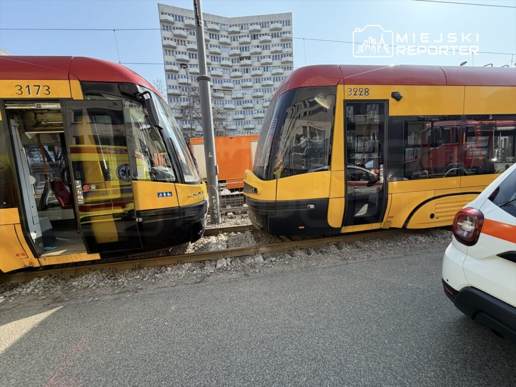 Dwa tramwaje w kolorze żółto-czerwonym stoją na torach obok siebie, w tle widać nowoczesny budynek mieszkalny.