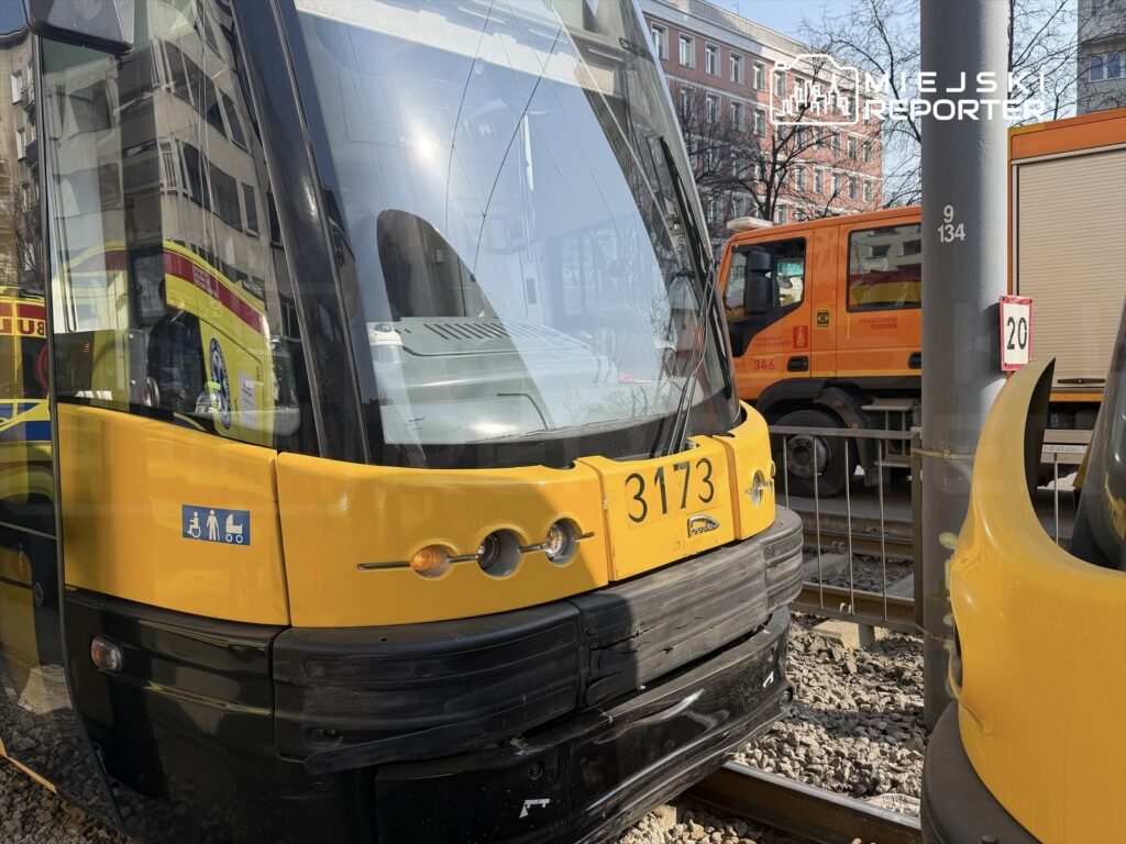 Żółty tramwaj z numerem 3173 zatrzymany na torowisku, w tle widoczny pomarańczowy samochód ciężarowy.