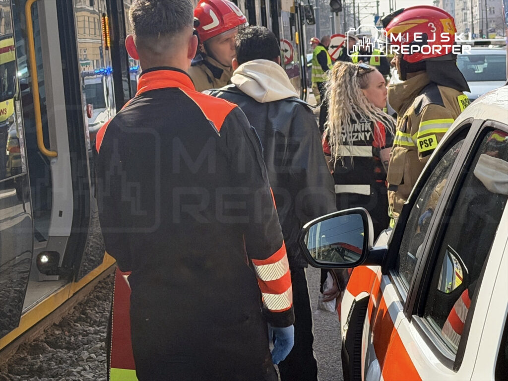 Grupa ratowników w odblaskowych strojach rozmawia z osobą poszkodowaną obok tramwaju na ruchliwej ulicy.