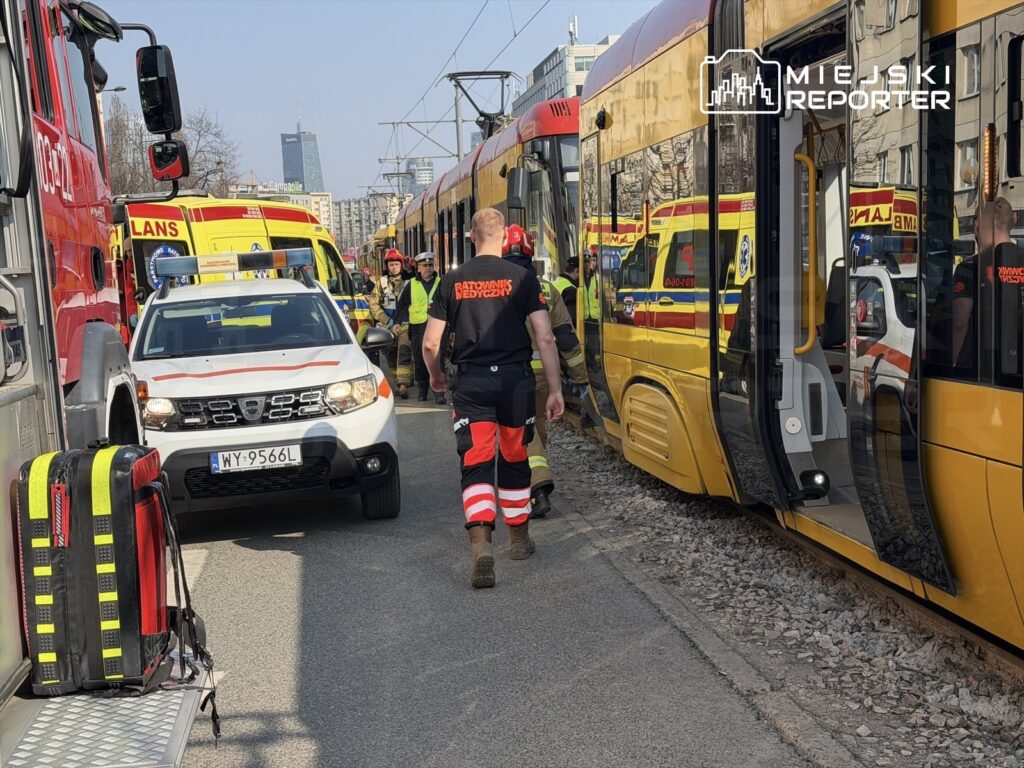 Ratownicy medyczni i strażacy pracują przy wypadku tramwajowym na miejskiej ulicy, obok żółtego tramwaju.