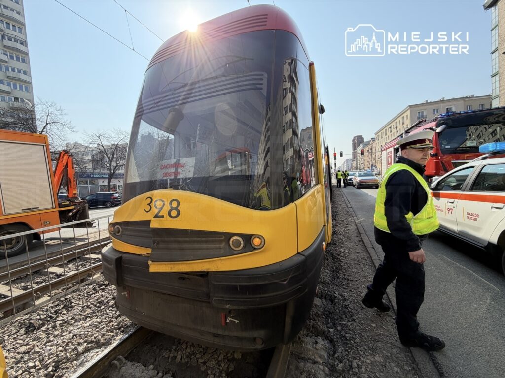 Tramwaj numer 328 zatrzymany na torach, obok policjant w kamizelce odblaskowej kierujący ruchem, w tle pojazdy ratunkowe.