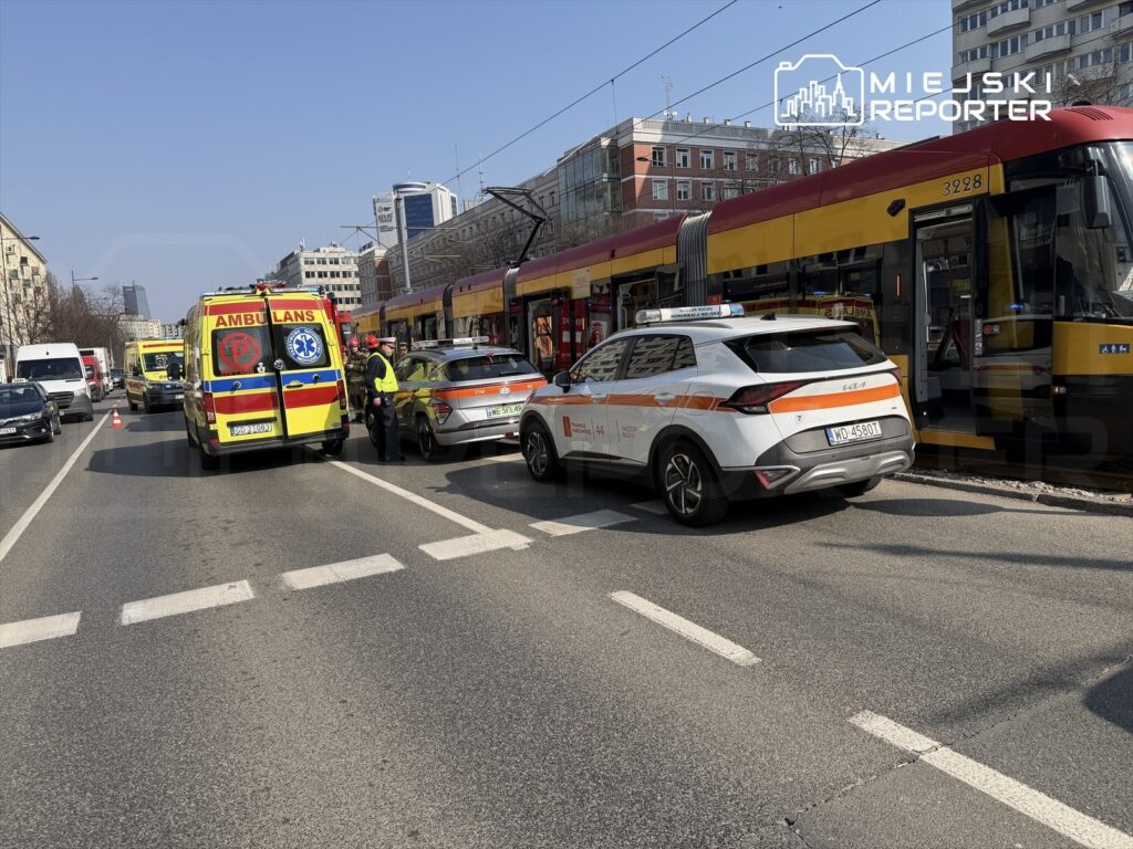 Karetka pogotowia oraz radiowóz policji na zatłoczonej ulicy, obok zatrzymanego tramwaju, w trakcie interwencji.