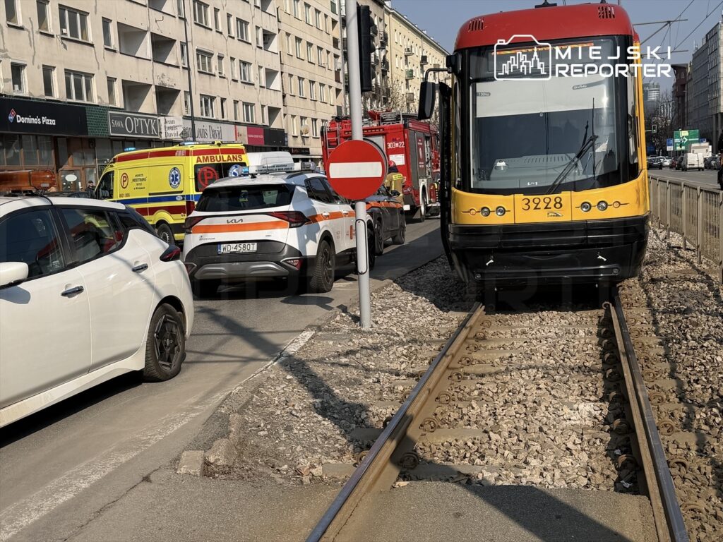 Tramwaj z numerem 3928 stoi na torach, obok pojazdy ratunkowe oraz policyjny samochód na ruchliwej ulicy.