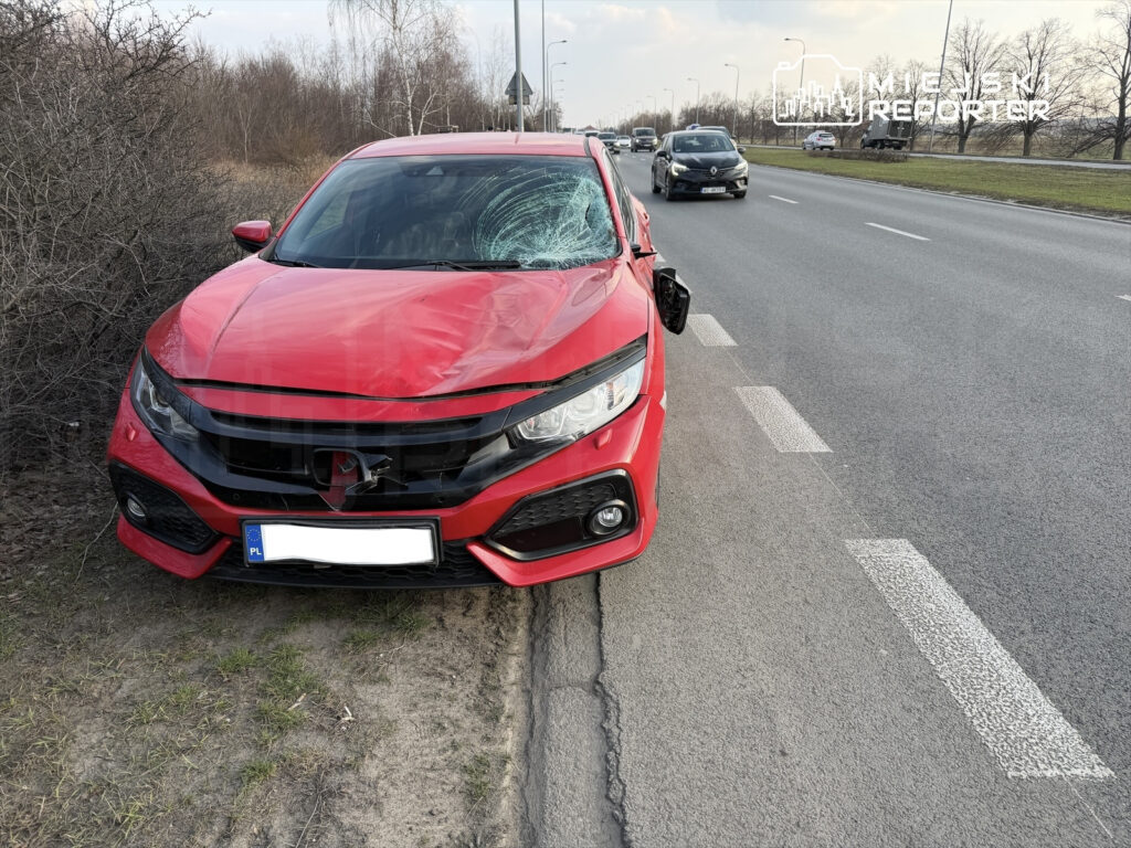 Czerwony samochód osobowy z uszkodzoną szybą stoi przy drodze, obok zarośli i przejeżdżających pojazdów.