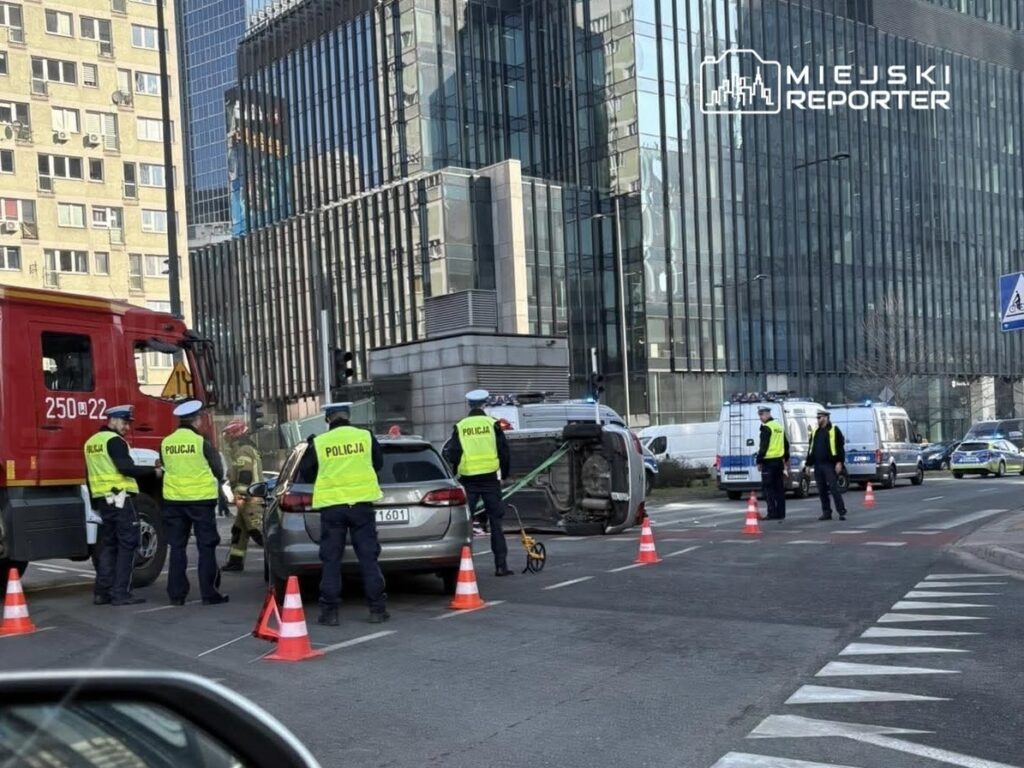 Grupa policjantów w kamizelkach odblaskowych zabezpiecza miejsce wypadku, gdzie przewrócone auto blokuje ruch.
