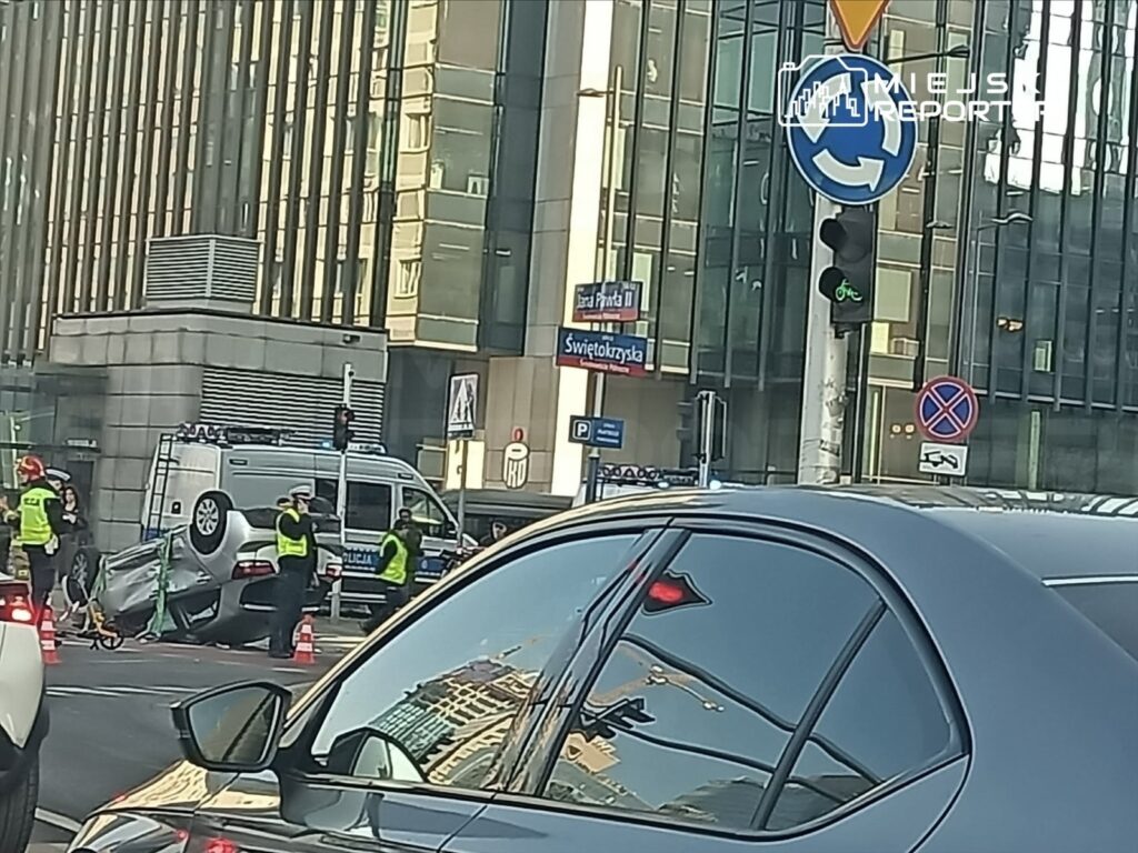 Na skrzyżowaniu stoją policjanci i ratownicy, obok przewrócone auto, a w tle nowoczesny biurowiec.