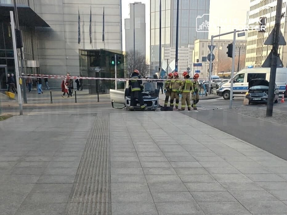 Grupa strażaków w hełmach bada uszkodzony samochód na ulicy przed nowoczesnym biurowcem.