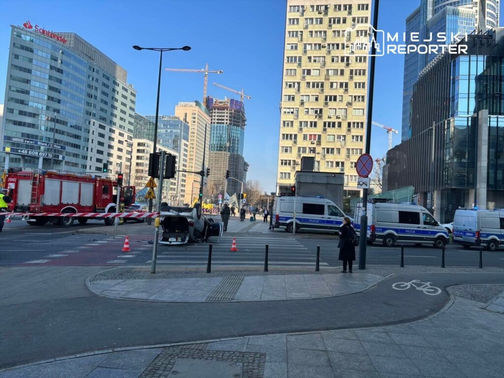 Na skrzyżowaniu w centrum miasta leży przewrócony samochód, obok stoją radiowozy i woz strażacki.