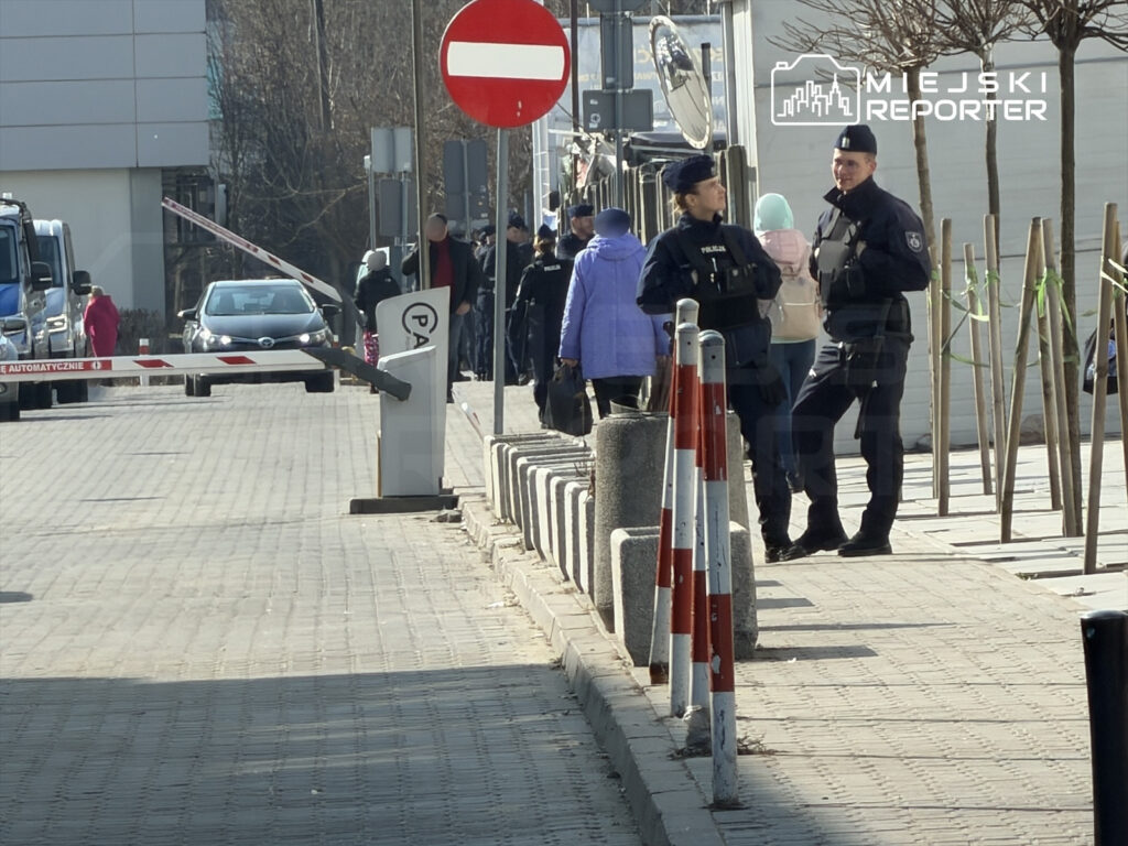 Grupa policjantów rozmawia na chodniku obok zamkniętej bramy wjazdowej, podczas gdy przechodnie mijają ich.