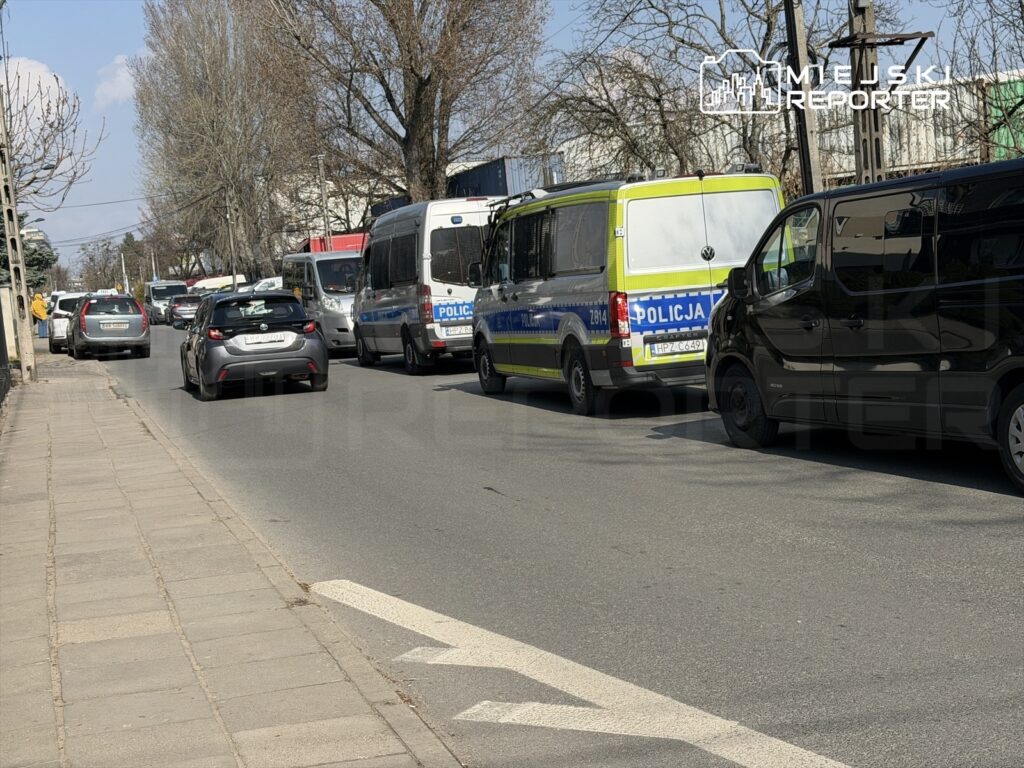 Ruchliwa ulica z samochodami w korku, w tym pojazdy policji oraz osobowe, w słoneczny dzień.