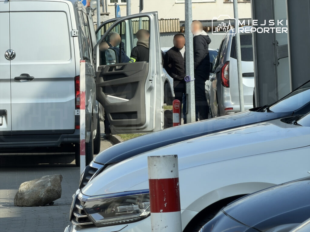 Grupa mężczyzn rozmawia obok otwartych drzwi białego pojazdu dostawczego na parkingu. W tle widać inne samochody.